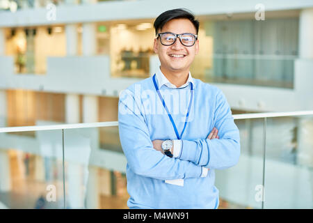 Waist up portrait of cheerful Asian businessman wearing glasses smiling happily at camera standing at glass balcony in modern office building, copy sp Stock Photo