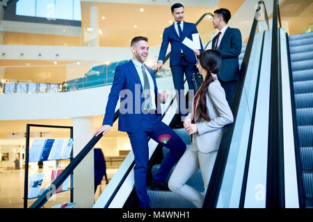Group of modern young business people riding escalator in office building and chatting cheerfully on the way, copy space Stock Photo