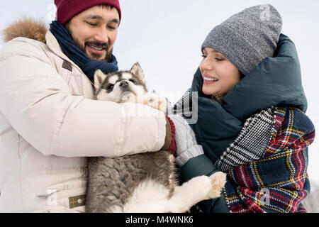 Portrait of modern young couple playing with cute husky puppy cuddling him outdoors on beautiful winter day Stock Photo