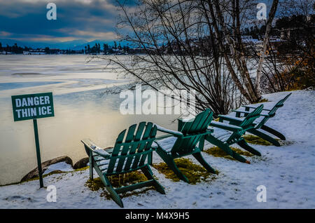 Posted sign warning of thin ice on Mirror Lake in Lake Placid, NY. Four green Adirondack chairs line the edge of the lake, next to white birch trees.  Stock Photo