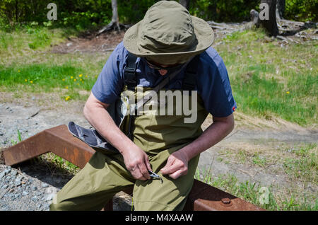 Lake Placid, NY, USA. A fisherman, fully attired in gaiters, sits on a rail to tie a lure. Stock Photo