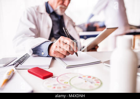 Unrecognizable senior doctor with tablet in his office. A nurse and a lying patient in the background. Stock Photo