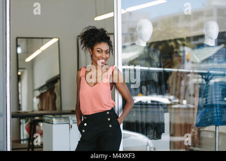 Smiling dress designer in her cloth shop with designer clothes on display. Woman entrepreneur standing at the entrance of her fashion studio. Stock Photo