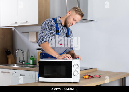 Happy Male Serviceman Using Screwdriver To Repair Microwave In Kitchen At Home Stock Photo