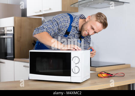 Happy Male Serviceman Using Screwdriver To Repair Microwave In Kitchen At Home Stock Photo