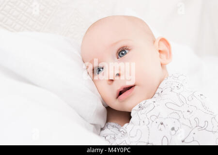 Adorable baby boy with blue eyes lying on a bed, looking away from camera. Cute toddler in pyjamas close up portrait. Stock Photo