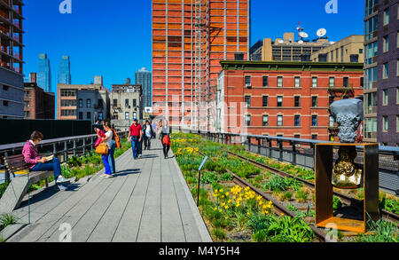 A golden sculpture, a woman sitting on a bench and reading a book, tourists taking photos, and pedestrians walking along path in High Line Park, NYC. Stock Photo
