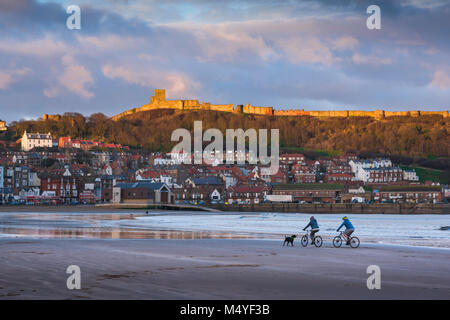 UK beach, out of season view of South Bay in Scarborough with the castle illuminated at sunset and a couple cycling on the beach,Yorkshire, England,UK Stock Photo