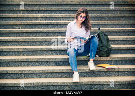 Beautiful girl, a student in glasses reads a book on the stairs. Education, training. Indian College Stock Photo