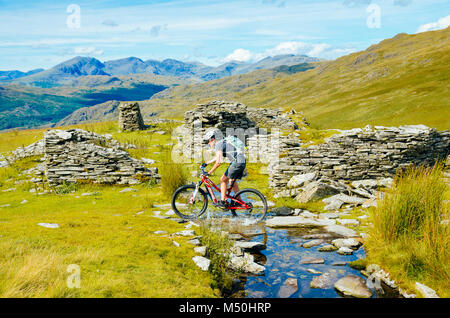 Female mountain biker passing quarry ruins at Walna Scar Quarries in the English Lake District with Scafell and Scafell Pike in the distance Stock Photo