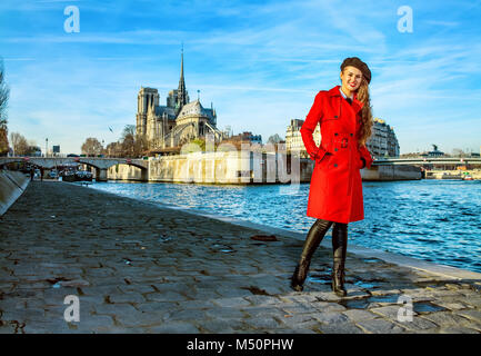 Bright in Paris. smiling elegant traveller woman in red trench coat standing on embankment near Notre Dame de Paris in Paris, France Stock Photo