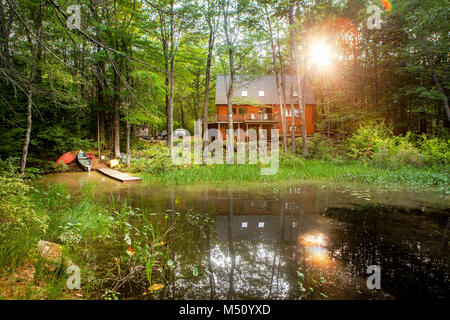 Morning sunshine bathes a modern log vacation home with a pond, dock and canoes in an idyllic setting surrounded by woodland, peace and quiet. Stock Photo