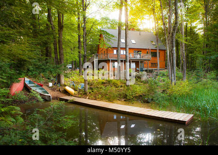 Morning sunshine bathes a modern log vacation home with a pond, dock and canoes in an idyllic setting surrounded by woodland, peace and quiet. Stock Photo