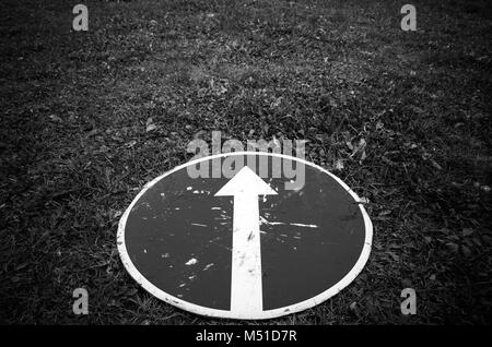 Ahead only, dark round road sign with white arrow lays on grass. Black and white photo Stock Photo