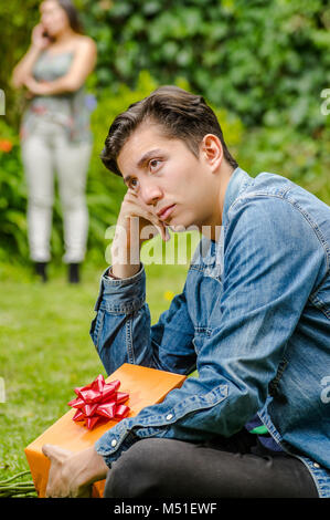 Close up of sad man wearing a jean jacket and black pants sitting in the ground holding gift with a blurred woman behind using her cellphone. Valentine's Day concept or friend zone concept Stock Photo