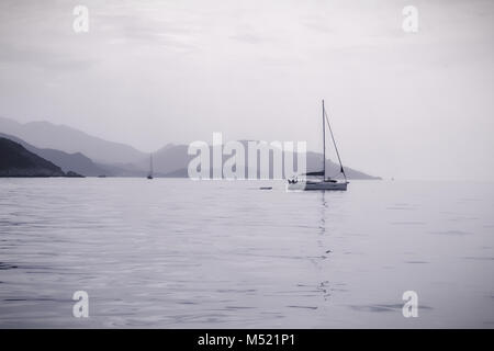 Sailing in calm seas with mountains in the distance in monotone dark grey to whiteout.  The mountains of Greece in the Mediterranean sea off Corfu Stock Photo