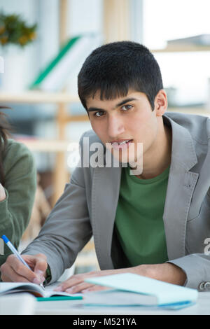 Handsome male student looking at camera while making notes sitting at classroom with other students Stock Photo