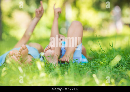 Group of happy unrecognizable children lying on green grass and pointing to the sky outdoors in summer park Stock Photo
