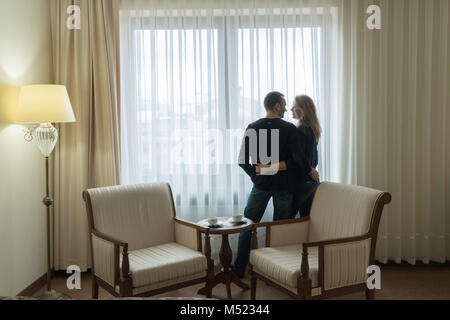 The newlyweds stand at the window of the hotel room and look at each other. Young family is very happy Stock Photo