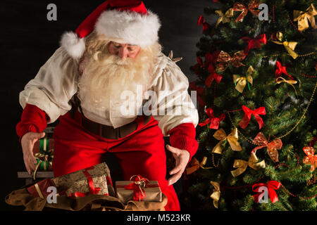 Santa Claus opening his sack and taking gifts under Christmas tree at night at living room at children's home Stock Photo