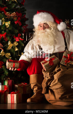 Santa putting gifts under Christmas tree in dark room Stock Photo