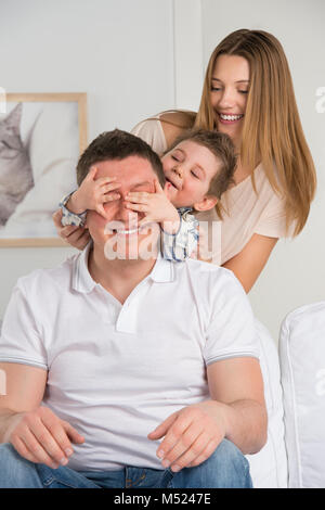 Happy family having fun in the living room: son surprisng his father by covering his eyes Stock Photo