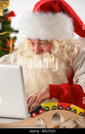 Real Santa Claus working on laptop with surprised face at home, surrounded by toys Stock Photo