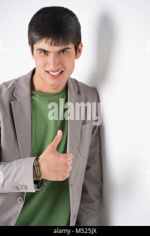 Happy smiling young man leaning against white wall and showing thumbs up Stock Photo