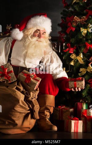 Santa putting gifts under Christmas tree in dark room Stock Photo
