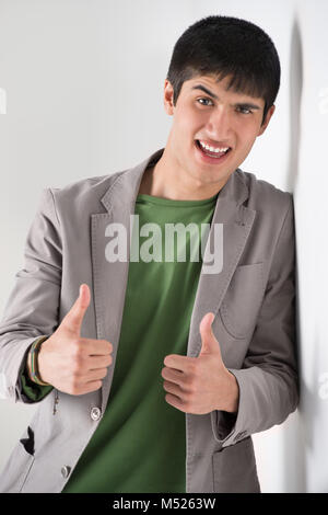 Happy smiling young man leaning against white wall and showing thumbs up Stock Photo