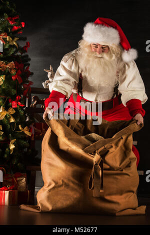 Santa Claus opening his sack and taking gifts under Christmas tree at night at living room at children's home Stock Photo