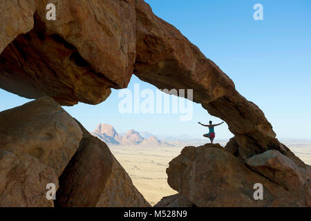 Woman doing yoga on rock formation in front of Spitzkoppe Mountain during bouldering expedition, Erongo region, Namibia Stock Photo