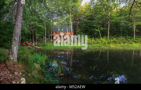 Morning sunshine bathes a modern log vacation home with a pond, dock and canoes in an idyllic setting surrounded by woodland, peace and quiet. Stock Photo