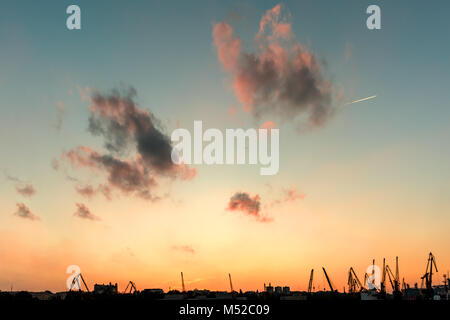 red gray clouds against a blue sky with a yellow orange sunset above the tower cranes buildings and structures of the evening port city Stock Photo
