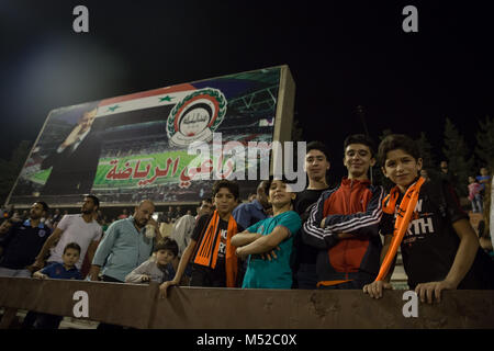 Boys pose in front of a billboard featuring Syrian President Bashar al Assad at the Syrian Football Cup final in Tishreen Stadium. The Syrian football cup final was played between Al-Wahda and Al-Karamah football teams. Despite ongoing conflict in the war torn country of Syria, the Syrian football cup final was hosted in the capital city of Damascus which is under the Syrian government control with many locals football fans attended the match. Stock Photo