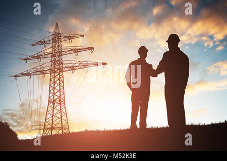 Silhouette Of Two Engineers Shaking Hands With Electricity Pylon Against Dramatic Sky Stock Photo