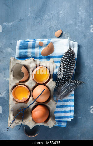 Carton tray with fresh eggs, whole and broken, feathers and tree branches on a checkered napkin. Top view with copy space. Spring, Easter concept. Stock Photo