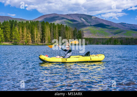 Middle aged woman having fun on the lake in the inflatable kayak; forest and mountains in the background; Colorado, USA Stock Photo