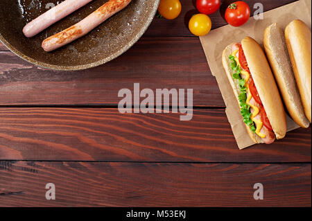 Hot Dog on craft paper bag with sausage in pan, bun, tomatoes and salad flat lay on wooden background. Top view. Stock Photo