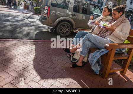 Two woman eat their lunch on a wooden public bench on the brick sidewalk by State Street in Santa Barbara, California. Stock Photo