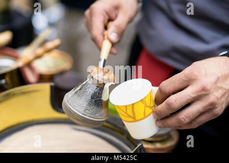 Close-up of hands of man making traditional turkish coffee in copper turk on hot sand and pours into disposable paper cup. Coffee arabic preparation concept Stock Photo