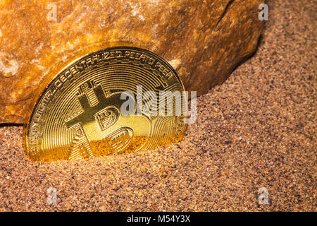 The gold coin of the crypto currency Bitcoin lies in the sand near the orange rock. Stock Photo