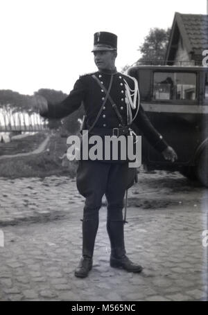 1920s, historical picture showing a young French policeman or 'gendarme' in his decorative military uniform, with tunic and breeches, standing on a village cobbled road using hand signals to direct motor vehicles of the era. The 'Gendarmerie Nationale is part of the French armed forces and has been since 1791. Stock Photo