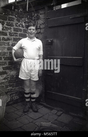 1920s, historical picture, shows a proud young boy standing in his football gear and boots holding a leather football in his backyard, England, UK. Stock Photo