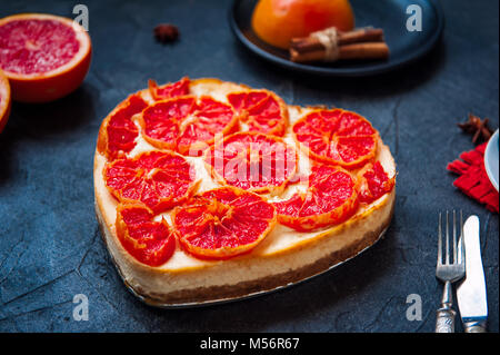 Baked cheesecake with red grapefruit slices in the shape of heart on the black stone background with ingredients and cutlery. Selective focus, space for text. Stock Photo