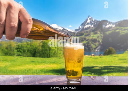 Hand of a man pouring beer from a bottle in a glass with a summer scenery in the background with mountains and green meadows, on a sunny day. Stock Photo