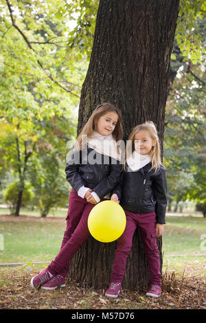 two daughters,little girls casually hang out by a beautiful old tree outdoors,smiling,autumn, park. Stock Photo