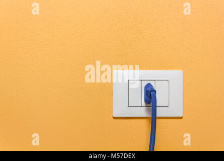 Blue plug inserted in a wall socket on an orange interior wall Stock Photo