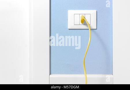 Yellow plug inserted in a wall socket on a light blue interior wall Stock Photo