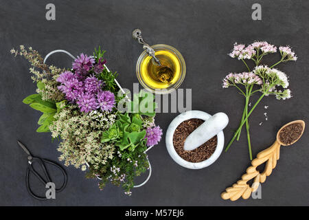 Herbal medicine apothecary preparation with fresh flowers and herbs, oil, scissors, valerian in a mortar with pestle and lovespoon on slate background Stock Photo